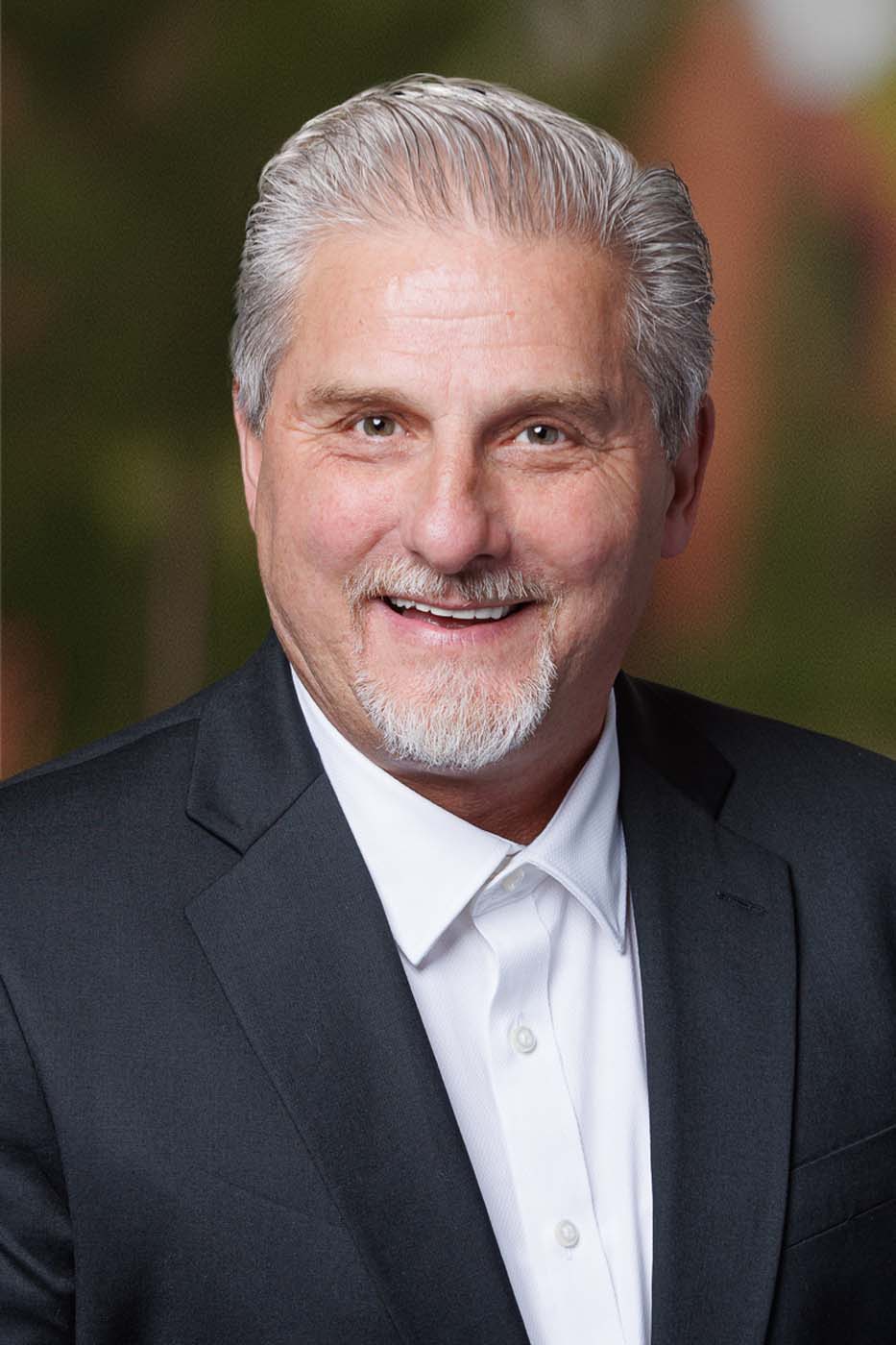 Smiling older man with gray hair and goatee, wearing a dark suit jacket and white shirt, posed in front of a blurred outdoor background—an experienced leader among Independent Cost Estimators.