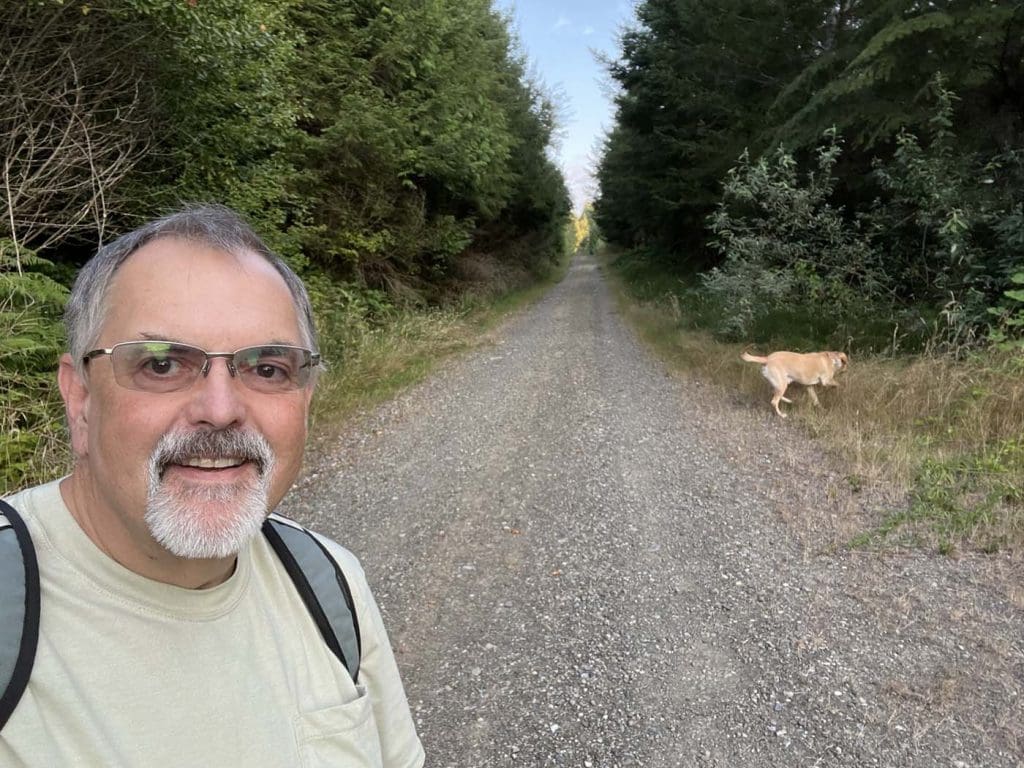 A man with glasses and a backpack takes a selfie on a gravel path lined with trees, while a yellow dog walks ahead on the right side.