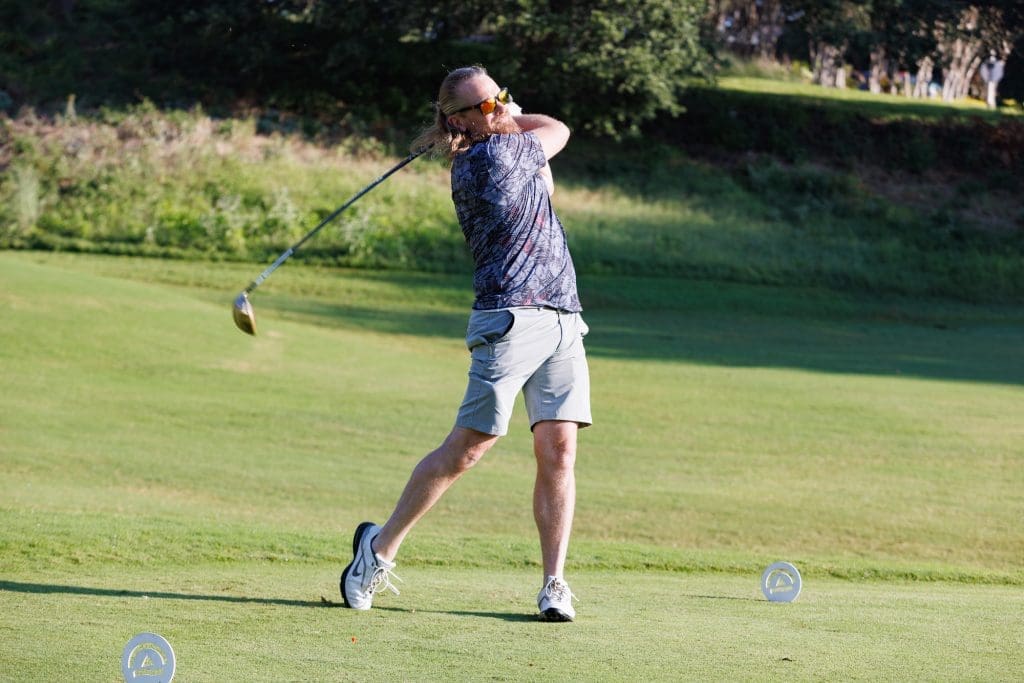 A person wearing a patterned shirt and shorts swings a golf club on a grassy golf course.