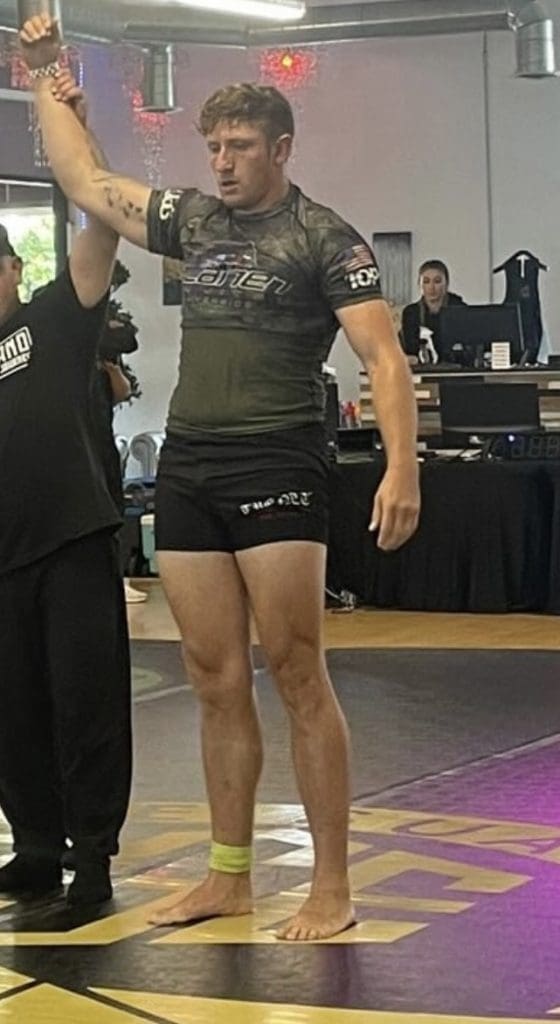 A barefoot male athlete stands on a mat as a referee raises his arm, signaling victory in a grappling or jiu-jitsu match.