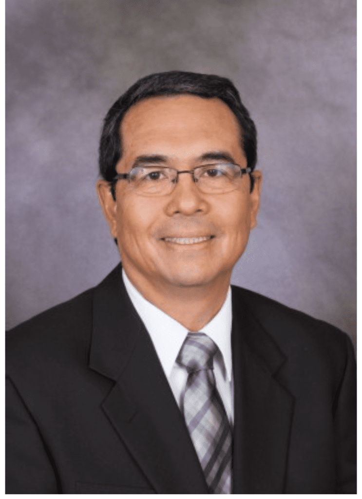 Man wearing glasses, a dark suit, white shirt, and plaid tie, smiling in a professional studio portrait with a gray background.