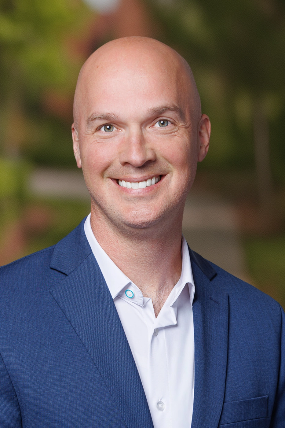 A bald man wearing a blue suit jacket and white shirt smiles at the camera, with a blurred outdoor background featuring greenery—reflecting the professionalism often found among Independent Cost Estimators.