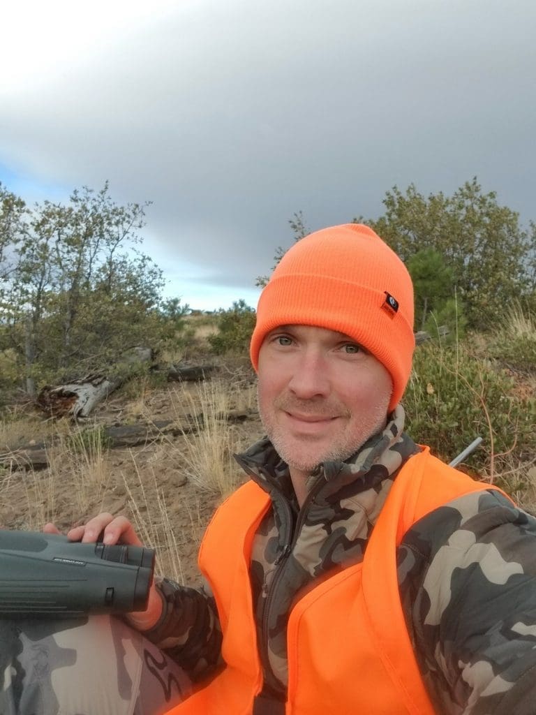 A man wearing an orange beanie and safety vest with camouflage clothing sits outdoors holding binoculars, with shrubs and cloudy sky in the background.