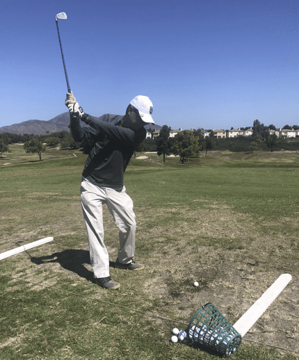 A golfer in a black long-sleeve shirt and white cap prepares to swing at a golf ball on a grassy driving range, with a basket of golf balls nearby.